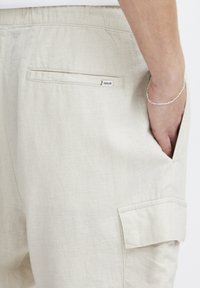 Crèmekleurige linnen broek met een achterzak met een label en een zij cargozak. De textuur lijkt zacht en ademend.