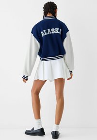 Marinblå och vit varsityjacka med "ALASKA" text på baksidan, ribbade ärmmuddar och kontrasterande ärmar, i kombination med en vit plisserad kjol.