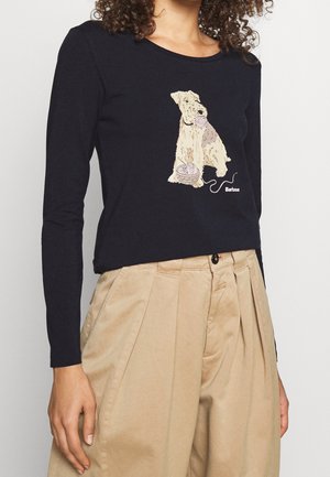 Žena v námořnicky modrém topu s dlouhým rukávem s ilustrací psa hrajícího si s klubkem příze, v kombinaci s béžovými plisovanými kalhotami s vysokým pasem.