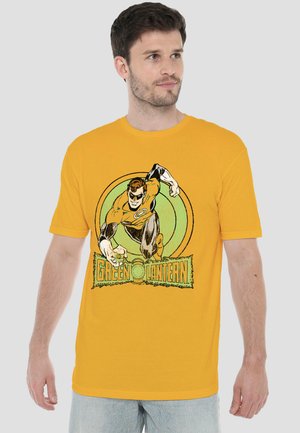 Mann trägt gelbes T-Shirt mit Green Lantern Superhelden-Grafik und Logo, schaut seitlich vor schlichtem Hintergrund.