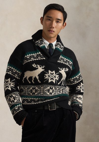 Pull noir en lainage avec motifs verts et crème, incluant des orignaux et des flocons de neige. Porté avec une chemise à col et un pantalon noir.