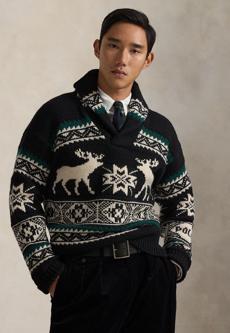Μαύρο πλεκτό πουλόβερ με πράσινα και εκρού σχέδια, συμπεριλαμβανομένων ταράνδων και νιφάδων χιονιού. Συνδυασμένο με πουκάμισο με γιακά και μαύρο παντελόνι.
