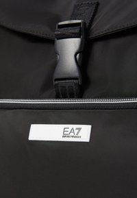 Black nylon backpack featuring a plastic buckle, silver zipper, and a white branded tag with 'EA7 Emporio Armani' logo. Soft texture.