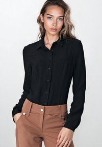 Czarna koszula z długim rękawem na guziki z klasycznym kołnierzykiem i gładką fakturą, w połączeniu z brązowymi spodniami szytymi na miarę z kontrastującymi guzikami.