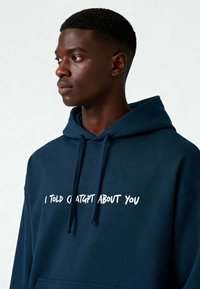 Navyblauwe hoodie van zacht materiaal, met een voorste kangoeroezak en witte tekst die zegt: "IK HEB CHATGPT OVER JOU VERTOELD."