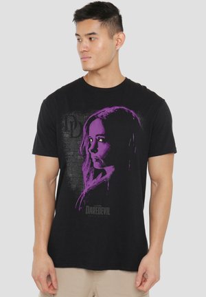 Young man wearing black Daredevil t-shirt with purple woman's face graphic, looking to his left against gray background.