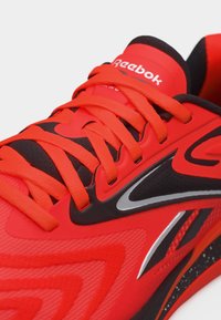 Κόκκινο και μαύρο αθλητικό παπούτσι Reebok με φωτεινά πορτοκαλί κορδόνια και ασημί λεπτομέρειες, που δείχνει μια κοντινή λήψη του πάνω μέρους και της περιοχής της γλώσσας.