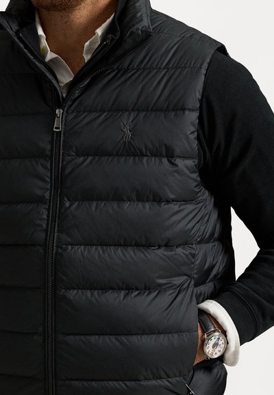Gilet rembourré noir avec un design matelassé horizontal, fermeture éclair, poches latérales et un petit logo sur la poitrine. Associé à un pull noir.