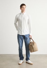 Camisa branca com padrões e botões, jeans azuis e ténis brancos. A segurar uma mala bege com detalhes castanhos. Fundo simples.