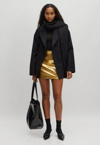 Zwarte oversized jas, zwarte turtleneck, metallic gouden minirok, zwarte handtas, nette knie-sokken, puntige zwarte schoenen, fijne texturen.