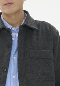 Temno siva volnena jakna s ovratnikom, ki ima na sprednji strani žepek in črne gumbe, pod katerimi je svetlo modra srajca z gumbi.