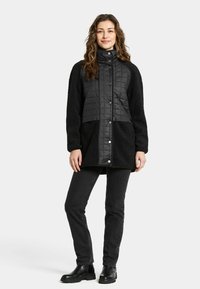 Kvinna med vågigt hår som bär en svart vadderad och fleecejacka, svarta jeans och svarta stövlar, stående mot en neutral bakgrund.