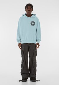 Lyseblå hættetrøje med frontlomme og cirkulært grafikdetalje; parret med sorte cargo-bukser med flere lommer.