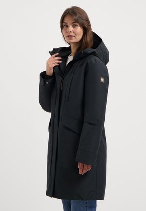 Femme portant un manteau d'hiver noir à capuche, avec de grandes poches avant, tenant le col, se tenant devant un fond clair.