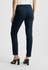 Pantalones slim-fit azul marino con una textura suave, que cuentan con dos bolsillos traseros y costuras decorativas. Combinados con sandalias negras de tacón cuadrado.