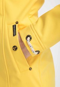 Gul vattentät jacka med en slät textur, prydd med flätad snodd och svarta, runda detaljer på ärmar och ficka.