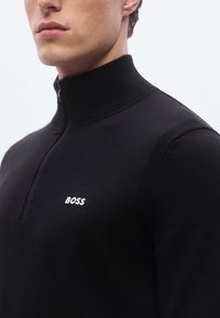 Svart genser med høy krage og halvlukking med glidelås. Har ribbet tekstur og hvitt "BOSS"-logo på venstre brystområde.