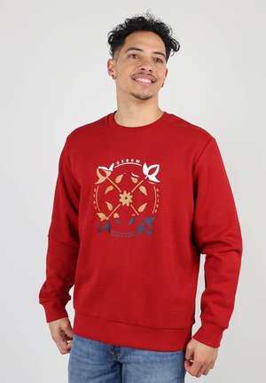 Sweat-shirt rouge à manches longues, avec un graphique circulaire en or, blanc et bleu marine, comportant des motifs floraux et de feuilles. Encolure ronde, poignets côtelés.