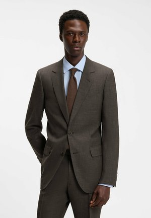 Uomo che indossa un completo marrone con camicia azzurro chiaro e cravatta marrone, in piedi davanti a uno sfondo bianco semplice, con una mano in tasca.