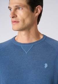 Maglione blu a collo rotondo realizzato in materiale morbido, con colletto e cuciture delle spalle a coste testurizzato, e un piccolo logo ricamato sul petto.