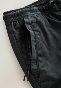 Schwarze leichte Hose mit einem elastischen Bund, Kordelzug und einer seitlichen Reißverschlusstasche. Der Stoff hat eine weiche, glatte Textur.