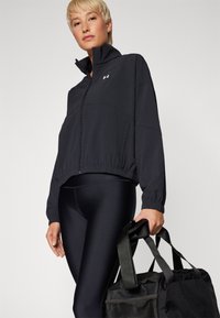 Chaqueta deportiva negra recortada con cremallera, cuello alto y puños elásticos, combinada con leggings negros y un bolso de viaje texturizado.