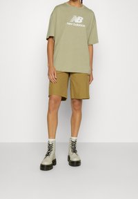 Khaki oversized tričko s bílým logem "New Balance", béžové kraťasy a světle šedé šněrovací boty s tlustou černou podrážkou.