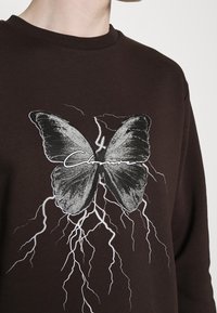 Sudadera marrón con un gráfico audaz de una mariposa negra, patrones de rayos blancos y una etiqueta con texto en escritura debajo de la mariposa.
