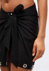 Κοντινό πλάνο μέσης με μαύρο σαρόνγκ δεμένο στη μέση πάνω από μαύρο μαγιό με πουά, ασημένιες οπές στο τελείωμα.