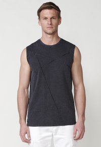 T-shirt sans manches gris foncé avec une surface texturée, présentant des motifs de couture en diagonale et un col rond, porté avec un pantalon blanc.