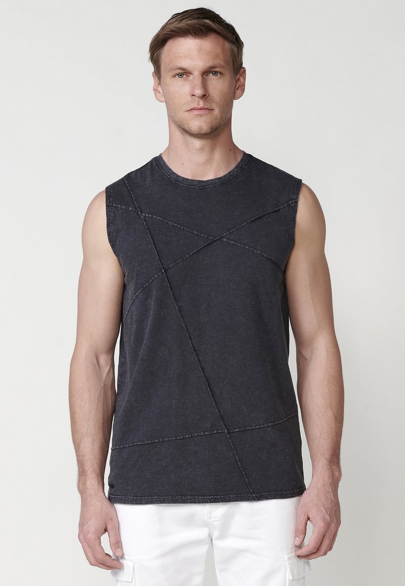 T-shirt sans manches gris foncé avec une surface texturée, présentant des motifs de couture en diagonale et un col rond, porté avec un pantalon blanc.
