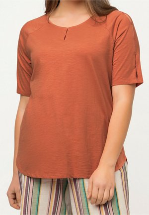 Femme portant un haut à manches courtes orange rouille avec une petite fente au niveau du col et un pantalon rayé multicolore, debout devant un fond uni.