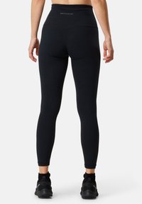 Leggings noirs taille haute en tissu lisse et extensible, dotés d'un logo au niveau de la ceinture arrière et d'une coupe ajustée, associés à des chaussures de sport noires.