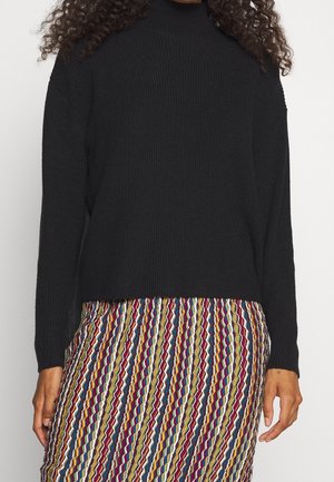 Personne portant un pull noir à manches longues associé à une jupe colorée à rayures verticales.