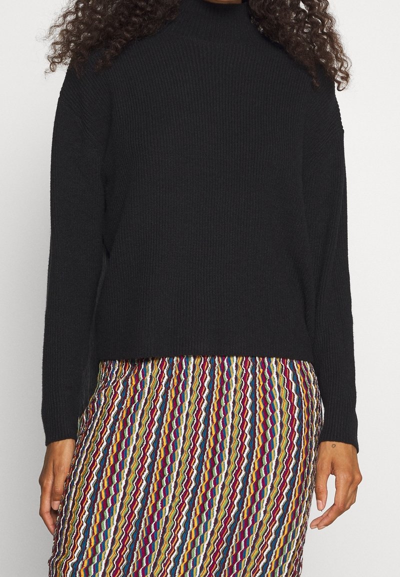 Personne portant un pull noir à manches longues associé à une jupe colorée à rayures verticales.