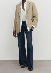 Blazer beige con spalle strutturate, camicia bianca con bottoni, canotta grigia, jeans in denim scuri a zampa d'elefante e scarpe nere con punta.  