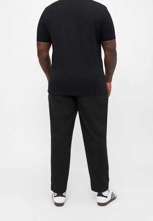 Mężczyzna z tatuażami na ramionach, ubrany w czarną koszulkę z krótkim rękawem, czarne spodnie, białe skarpetki i białe buty sportowe z czarnymi paskami, stojący tyłem.