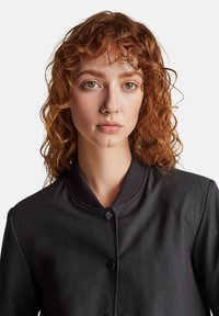 Giubbotto bomber grigio con colletto a coste, dotato di chiusura con bottone a pressione. Il tessuto ha una texture sottile ed è cucito per un look aderente.