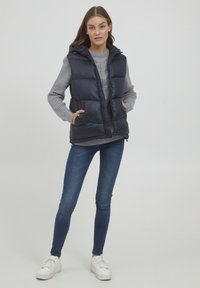 Marineblauer Puffer-Weste mit Kapuze, Reißverschluss und zwei Fronttaschen, getragen über einem hellgrauen Kabelstrickpullover und blauen Jeans.