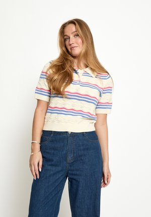 Kobieta o długich, jasnobrązowych włosach, ubrana w kremowy, dziergany polo z różowo-niebieskimi paskami oraz niebieskie jeansy z wysokim stanem na białym tle.