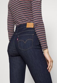 Mörka denimjeans med hög midja, med signatur bakfickor och en läderpatch med varumärkeslogotyp. Randig långärmad topp synlig.