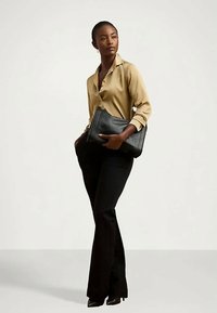 Ženska stoji, oblečena v bež satenasto bluzo, črne hlače, črne petke in drži veliko črno usnjeno torbico.