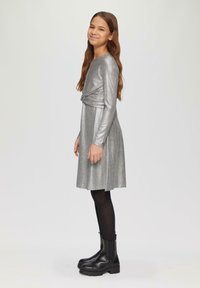 Silbernes Langarmkleid mit Wickeldesign, strukturiertem Stoff und schwingendem Rock. Kombiniert mit schwarzen Knöchelstiefeln und schwarzen Strumpfhosen.