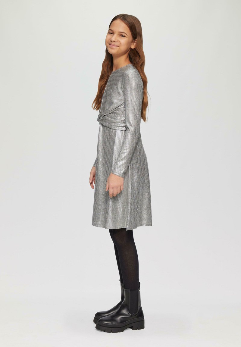 Silbernes Langarmkleid mit Wickeldesign, strukturiertem Stoff und schwingendem Rock. Kombiniert mit schwarzen Knöchelstiefeln und schwarzen Strumpfhosen.