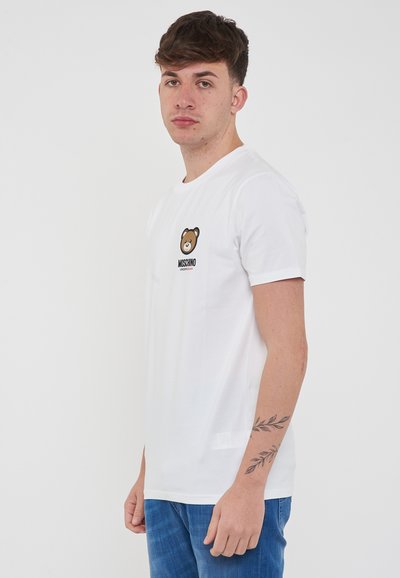 T-shirt bianca in cotone con grafica di un orso e testo "Moschino Underwear". Maniche corte e vestibilità standard. Modello che indossa jeans blu.