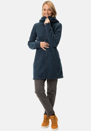 Femme souriante, portant un long sweat à capuche bleu foncé à fermeture éclair, un pantalon gris et des baskets orange-brun, debout sur un fond clair uni.