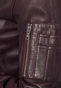 Ärmel aus burgunderrotem Lederjacke mit silbernem Reißverschluss und einer rechteckigen, gesteppten Tasche mit Nähdetails. Weiche Textur.