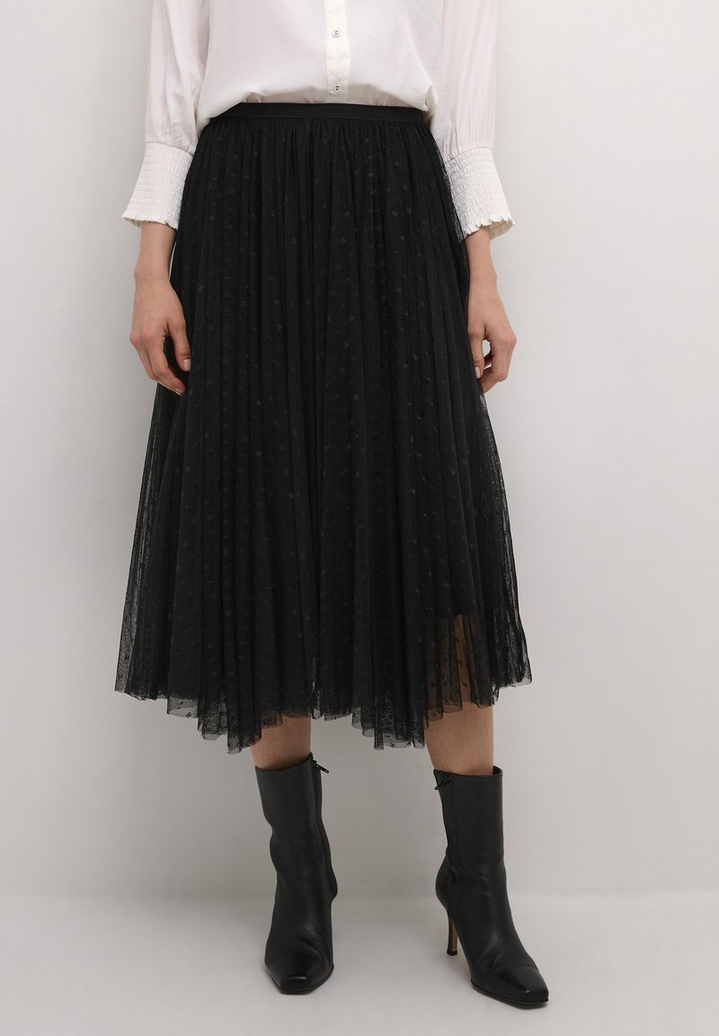 Black pleated tulle skirt with a textured dot pattern, high waist, layered design, paired with fitted black ankle boots.