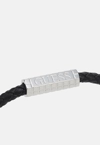 Bracciale in pelle intrecciata nera con una chiusura rettangolare in argento con il logo "GUESS" impresso su una superficie texturizzata.