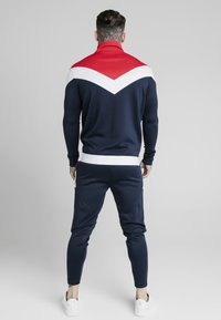 Granatowy dres z czerwono-białym wzorem w zygzak, dopasowanymi mankietami i elastycznym paskiem. Stylizowany z białymi sneakersami.